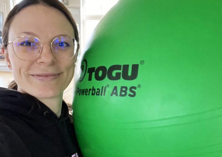 Frau mit Brille hält einen großen grünen Gymnastikball in der Hand.